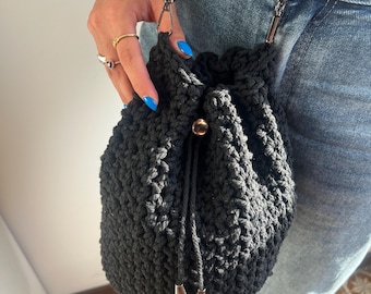 May include: A black, crocheted bucket bag with a drawstring closure and a silver chain strap. The bag has a textured appearance and a metal accent on the drawstring. The bag is held by a person wearing blue jeans.