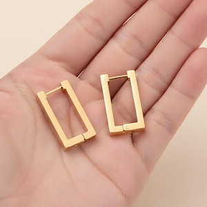 May include: A pair of gold-colored rectangular hoop earrings. The earrings are made of a shiny metal and have a simple, modern design. The earrings are shown on a light-colored background.