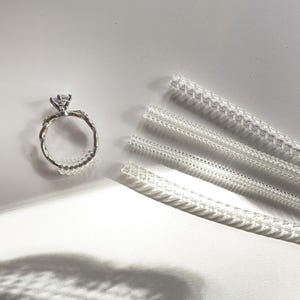 Puede incluir: Un anillo de compromiso plateado con una gema grande y transparente y una banda retorcida. También hay varios ajustadores de tamaño de anillo de plástico transparente. El anillo está sobre una superficie blanca.