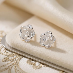 May include: A pair of silver stud earrings, each shaped like a rose. The earrings are presented on a cream-coloured surface, with a soft, blurred background. The rose design features detailed petals.