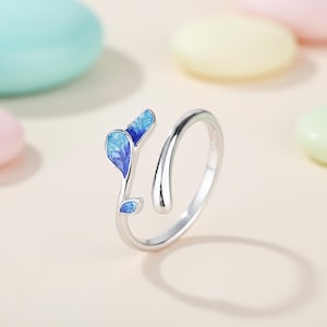 May include: A silver ring featuring a leaf design with blue enamel detailing. The leaves have a detailed, veined pattern in shades of blue. The ring is open, allowing for size adjustment. The background is a soft pastel colour.
