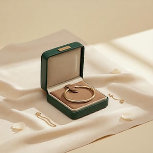 May include: An open, green jewelry box with a gold bracelet inside. The box sits on a cream-colored fabric, with a gold chain and a few white petals scattered around. The bracelet is a simple, elegant design.