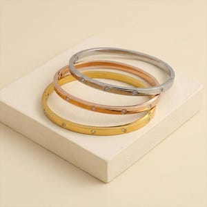 Könnte beinhalten: Drei Armreifen in Gold, Roségold und Silber. Jeder Armreif hat kleine, gleichmäßig verteilte, klare Edelsteine. Die Armreifen sind auf einer weißen, rechteckigen Oberfläche vor einem neutralen Hintergrund gestapelt.