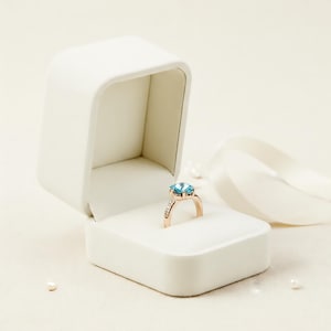Op de afbeelding: Een roségouden ring met een grote, lichtblauwe edelsteen wordt getoond in een open witte sieradendoos. De ring is gezet met kleinere, heldere stenen. Een wit lint en parels zijn op de achtergrond.