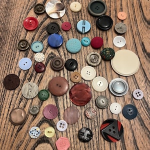 May include: A selection of buttons in diverse sizes, shapes, and hues, such as red, blue, green, and black. Some buttons feature multiple holes, while others have a shank. The buttons are displayed on a wooden surface.