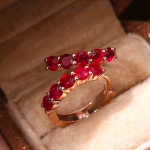 Pode incluir: Um anel de argola em ouro com design cruzado, apresentando duas filas de pedras preciosas redondas de um vermelho vibrante em cravação de garras. O anel tem um acabamento metálico polido e é exibido numa caixa de joias forrada a veludo.