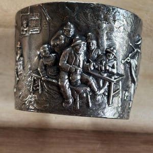 May include: A silver-toned metal napkin ring with a detailed relief scene of people gathered around a table. The relief depicts figures in period clothing, suggesting a historical or vintage theme. The ring is cylindrical and appears to be made of a solid metal.