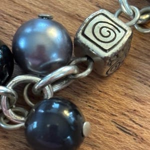 May include: Close-up of a silver-toned bracelet with a mix of beads and charms. The bracelet features a large, dark gray pearl, a black bead, and a square charm with a spiral design. The chain links are also silver.