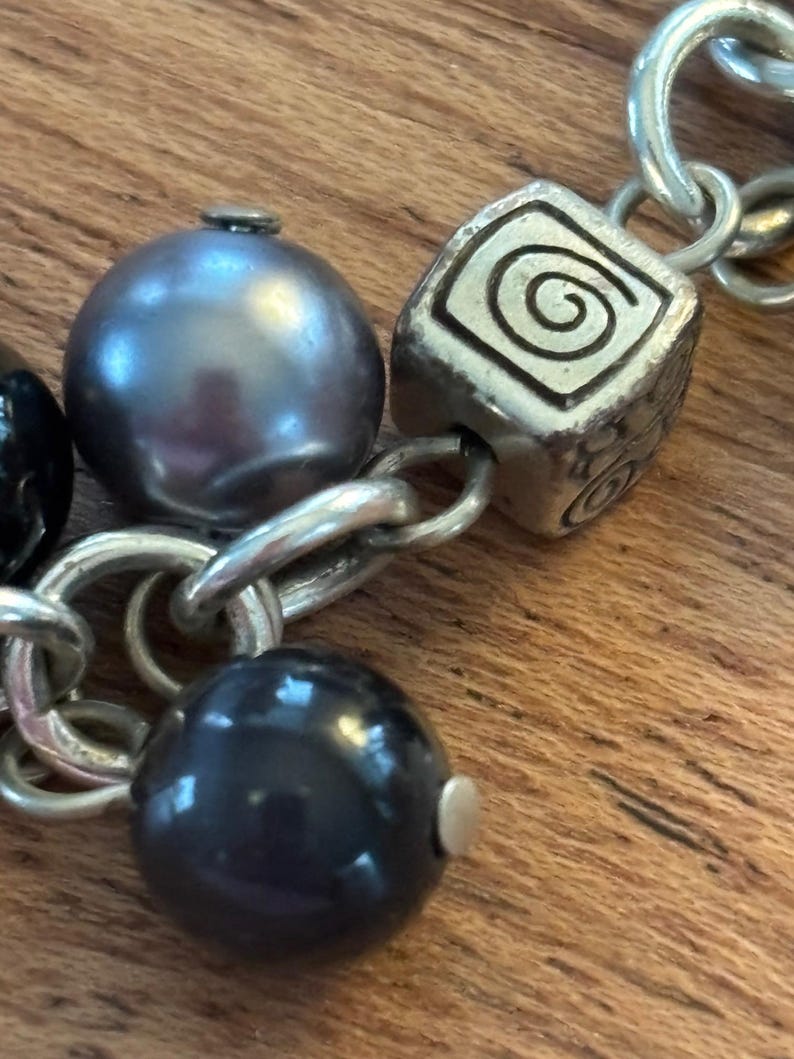May include: Close-up of a silver-toned bracelet with a mix of beads and charms. The bracelet features a large, dark gray pearl, a black bead, and a square charm with a spiral design. The chain links are also silver.