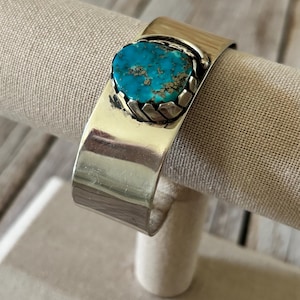 May include: A wide, polished silver cuff bracelet featuring a single, oval-shaped turquoise stone set in a serrated bezel. The bright blue stone displays natural brown matrix inclusions and is centered on the smooth, reflective metal band.