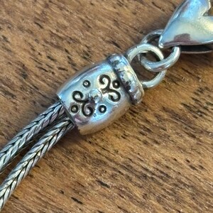 May include: Close-up of a silver-toned necklace clasp and chain. The clasp features an ornate barrel design with a decorative pattern. The chain is made of two strands of woven metal, and the clasp is connected to a heart-shaped element.