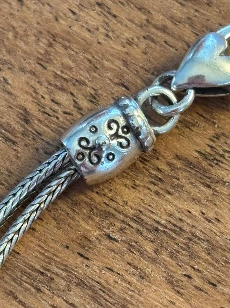 May include: Close-up of a silver-toned necklace clasp and chain. The clasp features an ornate barrel design with a decorative pattern. The chain is made of two strands of woven metal, and the clasp is connected to a heart-shaped element.