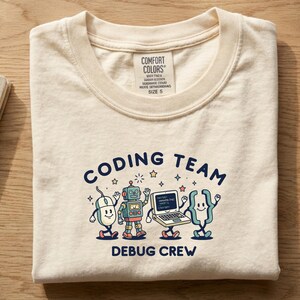 May include: A cream-coloured t-shirt with a graphic print featuring a computer mouse, a robot, a laptop, and a curly bracket character. The text reads CODING TEAM and DEBUG CREW. The shirt has a Comfort Colors brand label at the neck indicating size S.