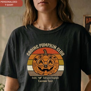 May include: A black t-shirt featuring a graphic of a jack-o'-lantern with glasses and coding symbols. The text reads "CODING PUMPKIN CLUB", "Emily", "Software Engineer", and "Custom Text". The design includes a sunset-style background.