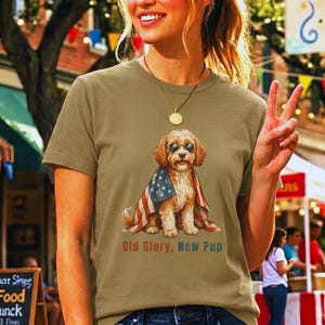 May include: A tan crew neck t-shirt featuring a graphic of a golden-colored dog wearing round sunglasses and draped in an American flag. The text below the dog reads Old Glory, New Pup in a mix of red and blue lettering on the front of the shirt.