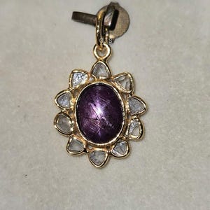 Puede incluir: Un colgante dorado con una piedra preciosa ovalada de color púrpura oscuro en el centro. La piedra preciosa está rodeada por un halo de piedras más pequeñas, claras y de forma irregular. El colgante está sujeto a un cierre dorado.