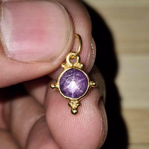 May include: A small gold pendant featuring a round, cabochon-cut purple star sapphire gemstone. The metal setting has a decorative, symmetrical design with small gold spheres at the top, bottom, and sides, and a circular bail at the very top.