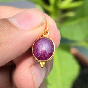 May include: A round, deep magenta cabochon gemstone pendant set in a polished gold-toned metal bezel. The pendant features a small gold-toned loop at the top for attachment and three tiny decorative gold-toned spheres at the bottom edge.