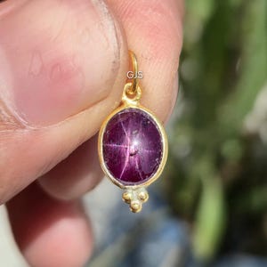 Pode incluir: Um pingente de ouro com uma pedra preciosa ovalada, vermelho escuro. A pedra preciosa tem um padrão em forma de estrela. O pingente é segurado por uma mão, com as letras "GJS" visíveis no laço de ouro.