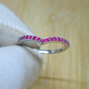 May include: A silver ring with a V-shaped design, set with small, vibrant pink gemstones. The ring is held against a white background. The letters "GJS" are visible on the ring.
