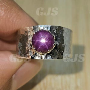 May include: A wide silver ring with a hammered texture. The ring features a large, round, deep-purple gemstone with a star-like pattern in the center. The gemstone is set in a gold-coloured bezel.