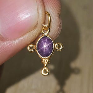 May include: A gold pendant featuring a purple star sapphire gemstone. The pendant has an oval-shaped stone with a star pattern, set in a gold frame with decorative accents. The pendant is held by a finger.