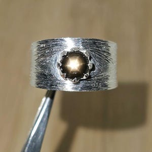 May include: A silver ring with a textured band and a central, star-shaped, dark brown gemstone. The ring is held by a pair of tweezers, and the initials "GJS" are visible on the ring's surface.