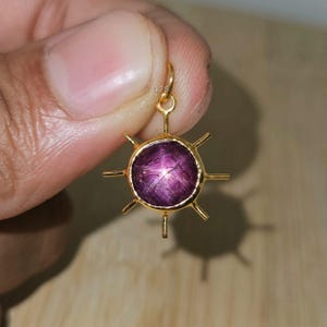 Puede incluir: Un colgante de oro con una piedra preciosa púrpura redonda en forma de estrella. El colgante tiene un diseño de sol con barras doradas radiantes. La piedra preciosa es de color púrpura intenso con un patrón en forma de estrella en el centro.
