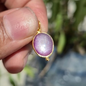 May include: A gold pendant featuring an oval, pale purple gemstone. The gemstone has a textured surface and is set in a gold bezel. The pendant is held by a finger against a blurred green backdrop.