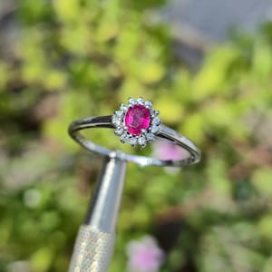 May include: A silver-toned ring featuring a central oval-cut vibrant pink gemstone surrounded by a halo of small, clear, round-cut stones. The band is thin and polished, with the letters GJS engraved on the inner surface of the metal setting.