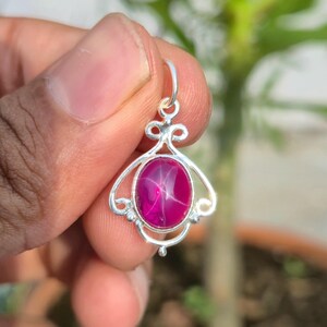 May include: A silver pendant featuring a central oval-shaped magenta star ruby gemstone. The metal setting has an intricate openwork design with decorative swirls and a small circular bail at the top for attaching to a necklace chain.