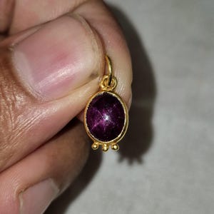 May include: A gold pendant featuring an oval-shaped, deep purple cabochon gemstone set in a smooth bezel. The base of the pendant is decorated with three small gold spheres, and a small gold jump ring is attached to the top for suspension.