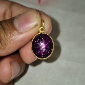 May include: A round pendant featuring a deep purple, faceted gemstone set in a polished gold-toned metal bezel. The pendant includes a small, circular gold-toned bail at the top for attachment, showcasing a rich, dark violet hue with subtle internal texture.