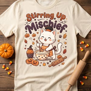 May include: A cream-colored t-shirt featuring a graphic of a white cat wearing a chef hat and apron, stirring a bowl. The text reads Stirring Up Mischief. Surrounding the cat are illustrations of pumpkins, a cupcake, a rolling pin, and candy corn.
