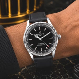 Könnte beinhalten: Eine silberfarbene Armbanduhr mit schwarzem Zifferblatt, auf dem der Text Breitling 1884, 17 Jewels und Shock Proof steht. Die Uhr hat eine geriffelte Lünette, silberne Zeiger und ein schwarzes Lederarmband. Das Zifferblatt enthält eine Minutenskala.