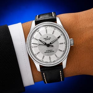 Könnte beinhalten: Eine Breitling Geneve Armbanduhr mit silbernem Sonnenschliff-Zifferblatt, schwarzen Stundenmarkierungen und silbernen Zeigern. Die Uhr verfügt über eine 24-Stunden-Skala, eine geriffelte Lünette und ein schwarzes Lederarmband mit weißen Nähten. Auf dem Zifferblatt steht Breitling Geneve, 17 Jewels und Shock Proof.