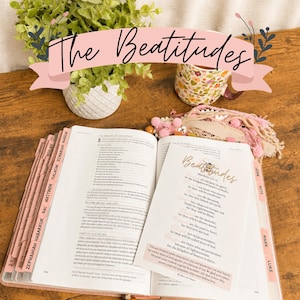 Puede incluir: Una Biblia abierta con pestañas rosas y una tarjeta impresa titulada The Beatitudes Matthew 5:3-12 sobre las páginas. Una taza con estampado floral y una pequeña planta verde en una maceta blanca texturizada se ven detrás del libro.