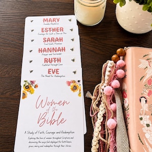 May include: A set of white cards titled Women of the Bible, featuring floral illustrations and bee motifs. The cards list names including Mary, Esther, Sarah, Hannah, Ruth, and Eve, each with a short phrase. A pink book with tassels and a candle are nearby.