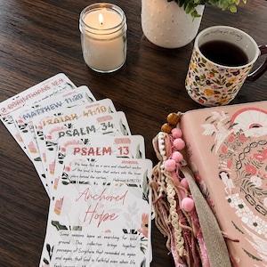 Op de afbeelding: Een stapel rechthoekige kaarten met bloemenillustraties en bijbelverzen, waaronder de tekst Anchored in Hope en Hebreeën 6:19. Naast de kaarten ligt een roze bijbel met decoratieve kwastjes, een brandende kaars in een glas en een bloemenmok.