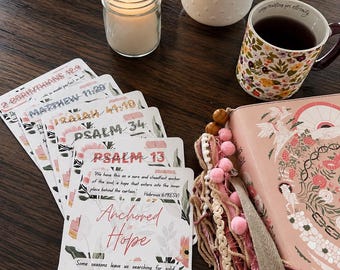 May include: A stack of rectangular cards featuring floral illustrations and Bible verses, including text like Anchored in Hope and Hebrews 6:19. Beside the cards is a pink Bible with decorative tassels, a lit candle in a glass jar, and a floral mug.