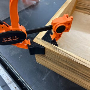 May include: An orange Jorgensen E-Z Hold clamp securing the corner of a wooden box. The box is made of light-colored wood with dark grain patterns. Another orange clamp is visible in the background, along with a black surface.