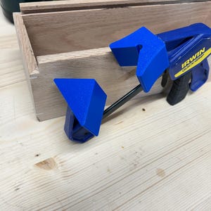 May include: A wooden box is held together by two blue and black Irwin Quick-Grip clamps. The clamps are securing the box's sides. The box is made of light-colored wood, and the clamps are made of plastic and metal.