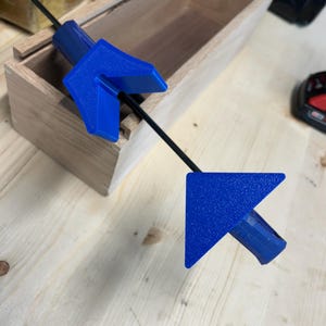 May include: A blue arrow-shaped tool with a textured surface, connected by a black rod, positioned near a wooden box. The tool's design features a right-angle shape, suggesting a measuring or alignment function.