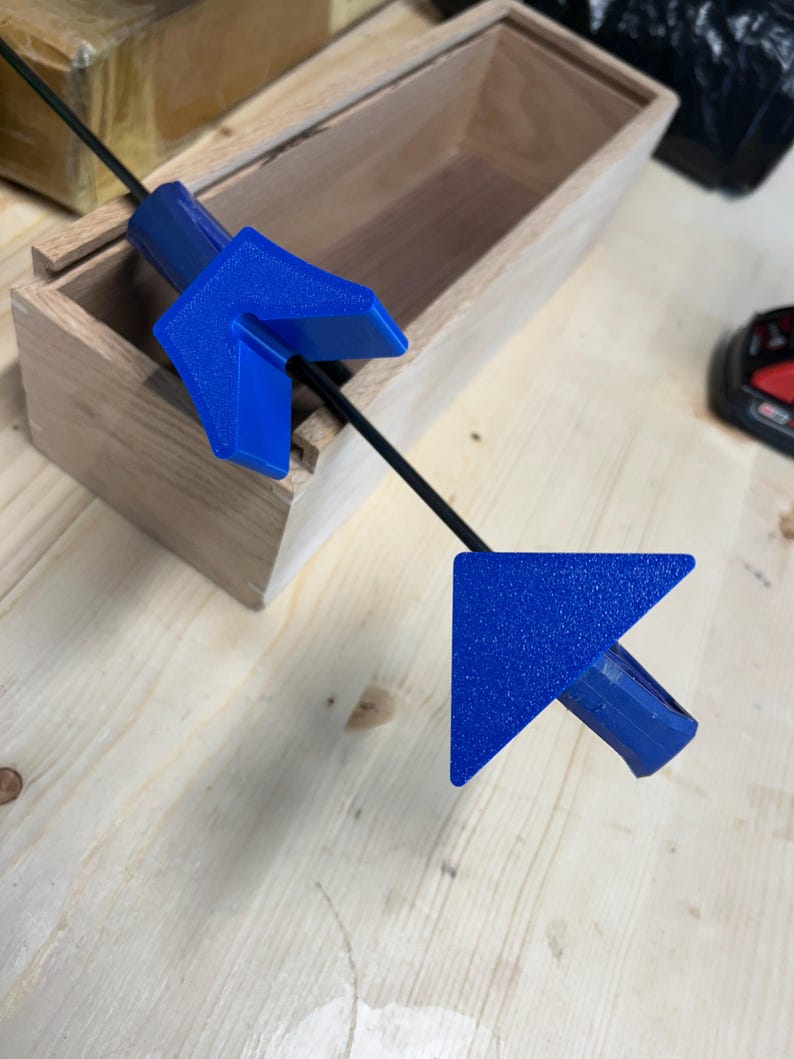 May include: A blue arrow-shaped tool with a textured surface, connected by a black rod, positioned near a wooden box. The tool's design features a right-angle shape, suggesting a measuring or alignment function.