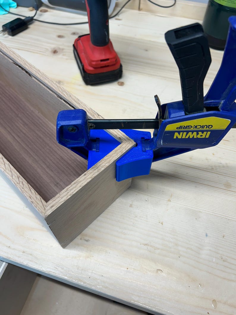 May include: A wooden box under construction, with a light-colored exterior and a darker interior. A blue and black Irwin Quick-Grip clamp secures the corner. A red power drill is visible in the background.