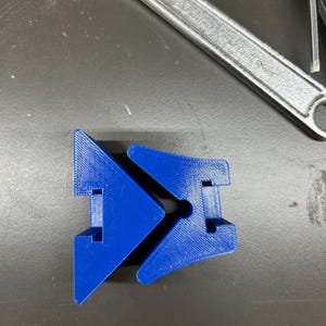 May include: A bright blue, 3D-printed plastic object with a unique geometric design. The object has a central hole and two angled sections. A silver adjustable wrench is in the background.