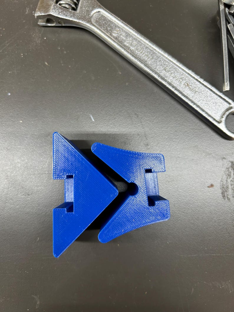 May include: A bright blue, 3D-printed plastic object with a unique geometric design. The object has a central hole and two angled sections. A silver adjustable wrench is in the background.