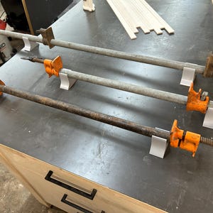 Könnte beinhalten: Drei Rohrspanner mit orangefarbenen und schwarzen Komponenten sind auf einer dunkelgrauen Werkbank angeordnet. Die Spanner werden für Holzbearbeitungs- und Bauprojekte verwendet. Die Werkbank hat eine helle Holzschrankbasis.