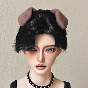 May include: A doll's head with black hair and brown dog ears, wearing a black choker necklace with a silver star pendant. The doll is on a black stand, against a white background.