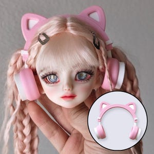 May include: A doll's head with light pink hair and blue eyes, wearing pink cat ear headphones with white accents. A smaller image of the headphones is in the lower right corner.
